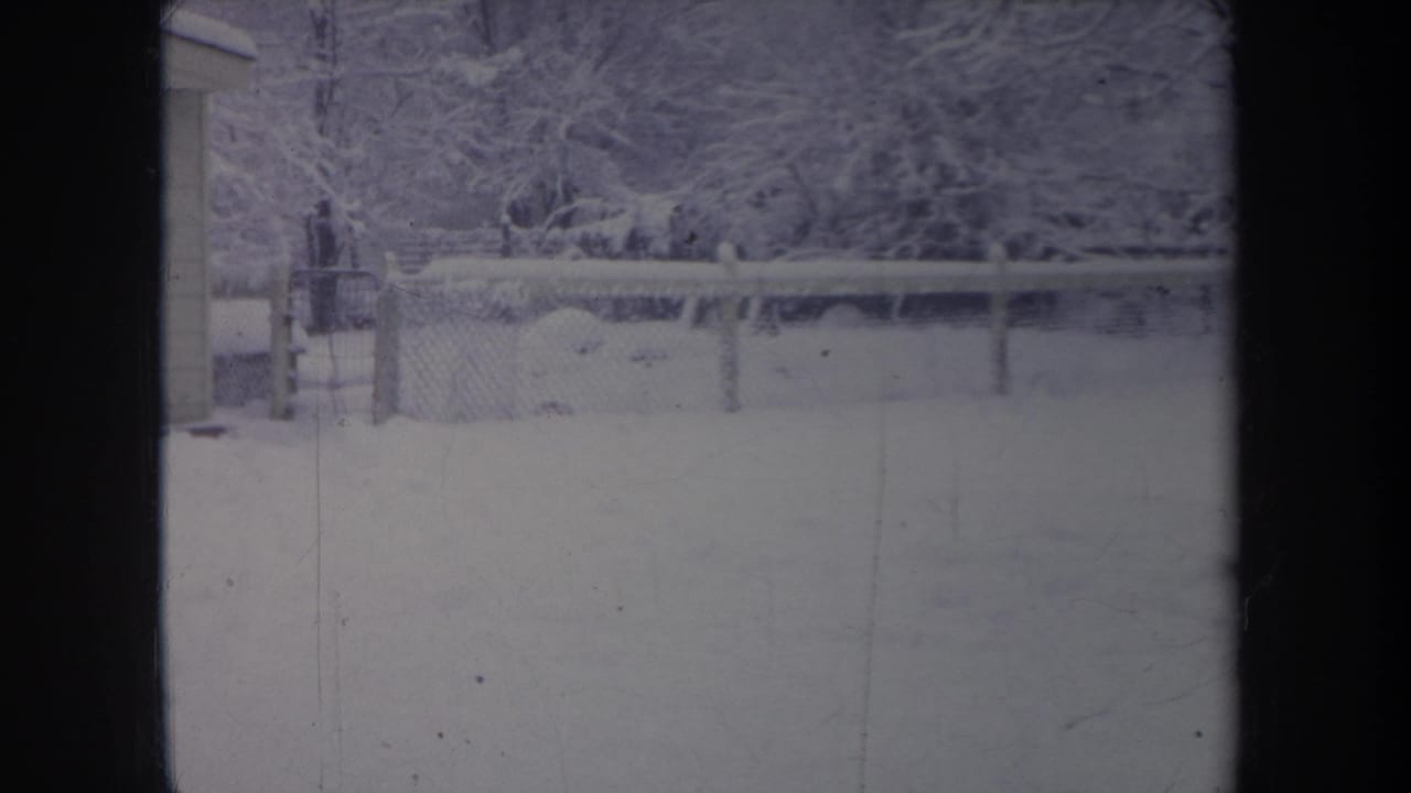 1968 Michigan winter frame with a chain-link fence, snow-covered yard, trees, and the edge of a small shed.