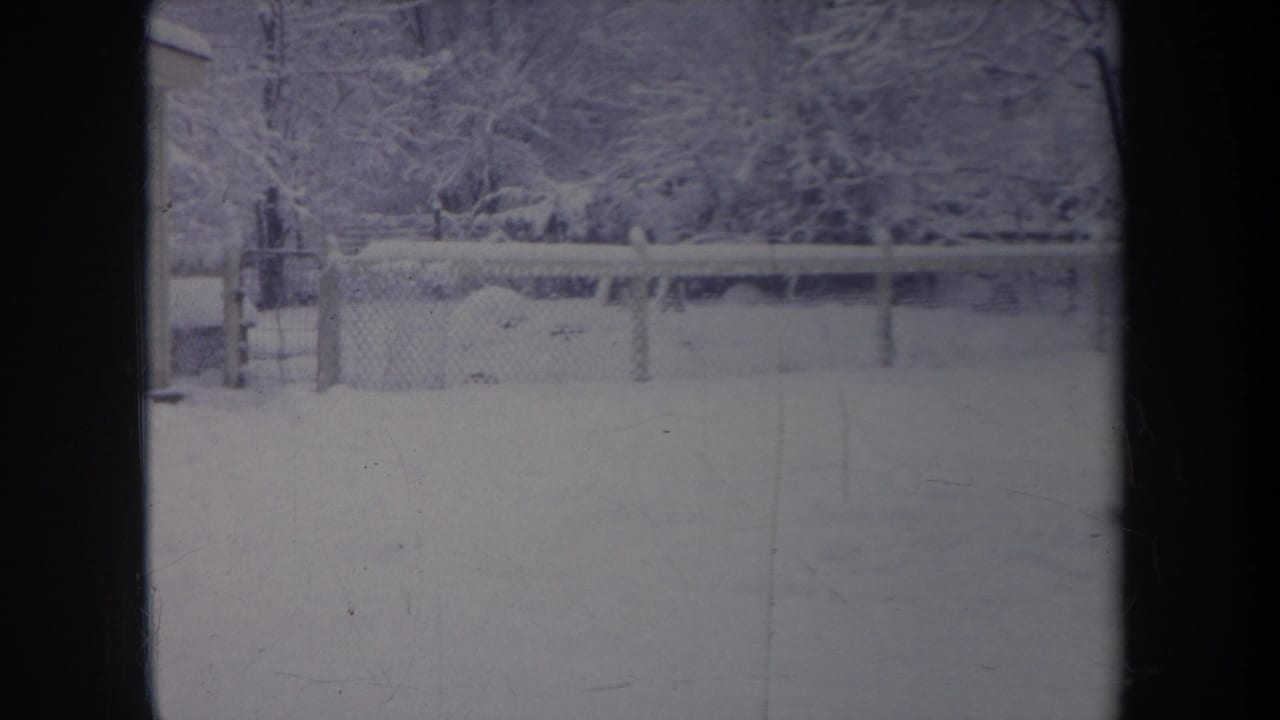 1968 Michigan 8mm frame showing a snow-covered yard, chain-link fence, and bare trees viewed through a dark window frame.