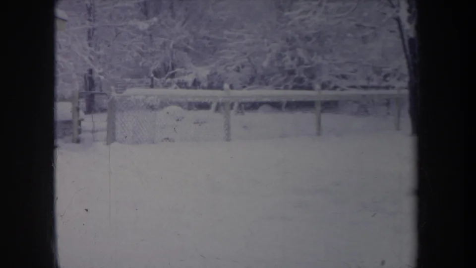 1968 Michigan winter home-movie frame with snow on the ground, a chain-link fence, and trees behind a dark window divider.