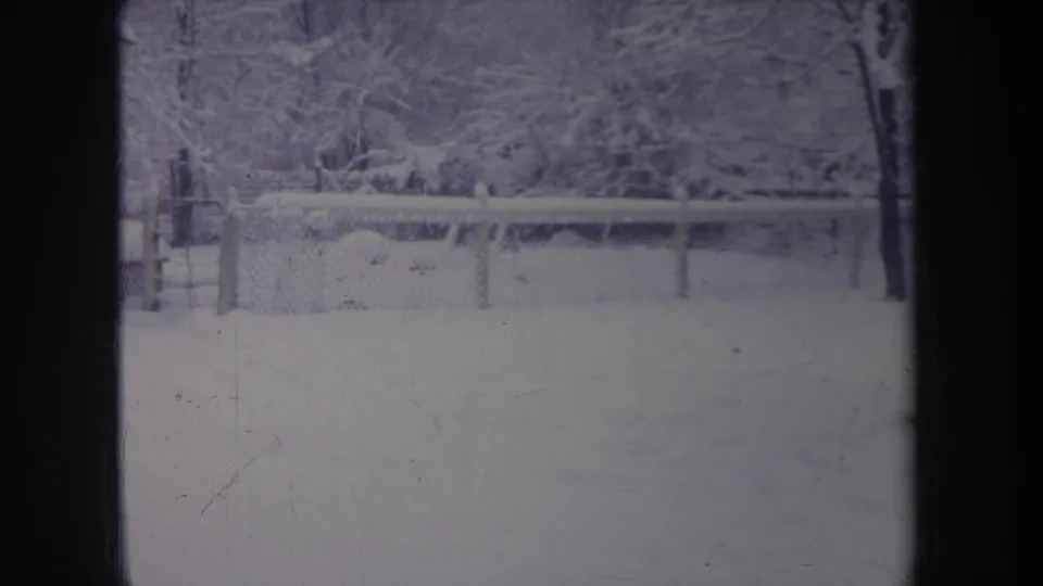 1968 Michigan 8mm still of a snow-covered residential yard, chain-link fence, and bare trees seen through a window opening.