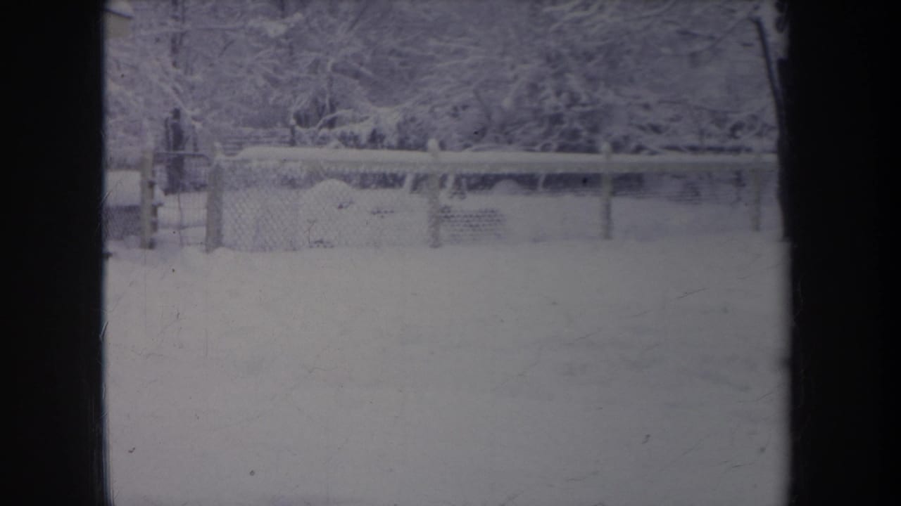 1968 Michigan home-movie frame showing a snowy backyard, chain-link fence, and tree line with a dark window frame in front.