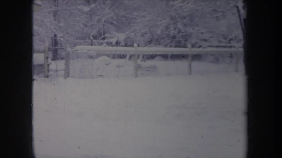 1968 Michigan home-movie frame showing a snowy backyard, chain-link fence, and tree line with a dark window frame in front.
