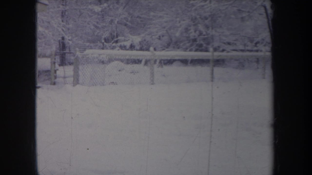 1968 Michigan 8mm frame showing a snow-covered backyard, chain-link fence, and trees under a faded blue winter cast.