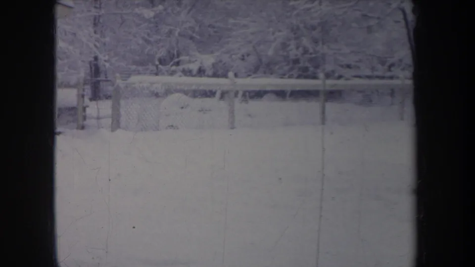 1968 Michigan 8mm frame showing a snow-covered backyard, chain-link fence, and trees under a faded blue winter cast.