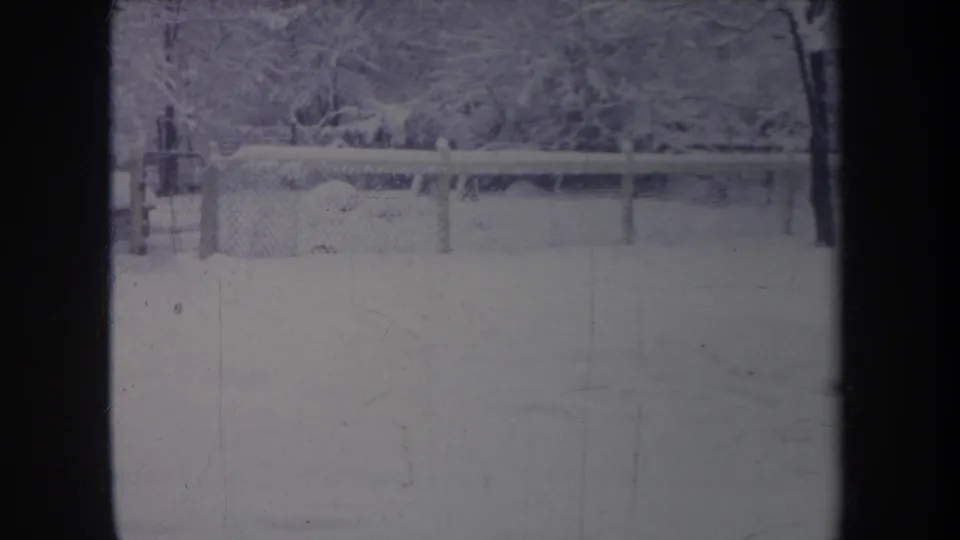 1968 Michigan home-movie still of a quiet snow-covered yard, fence, and tree line with visible 8mm wear.