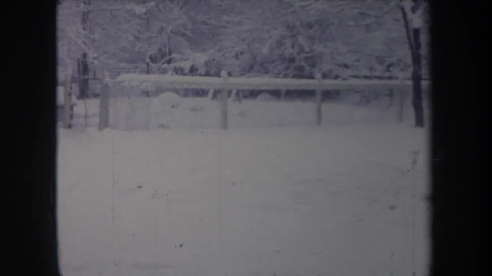 1968 Michigan home-movie frame showing a wintry backyard, chain-link fence, and snow-laden trees in faded 8mm color.
