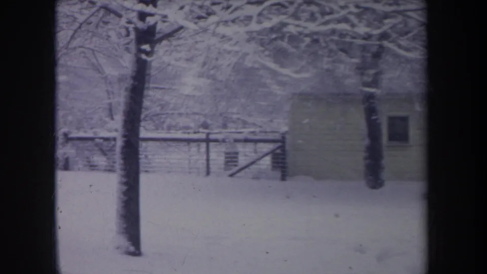 1968 Michigan home-movie frame showing a snow-covered yard, small shed, fence, and bare trees in soft 8mm color.