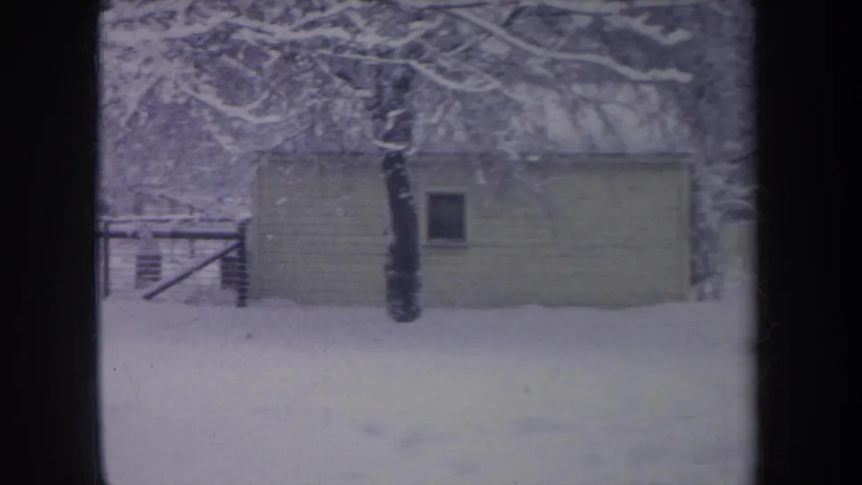 1968 Michigan 8mm frame of a small outbuilding in deep snow, with a tree trunk and fence behind it.