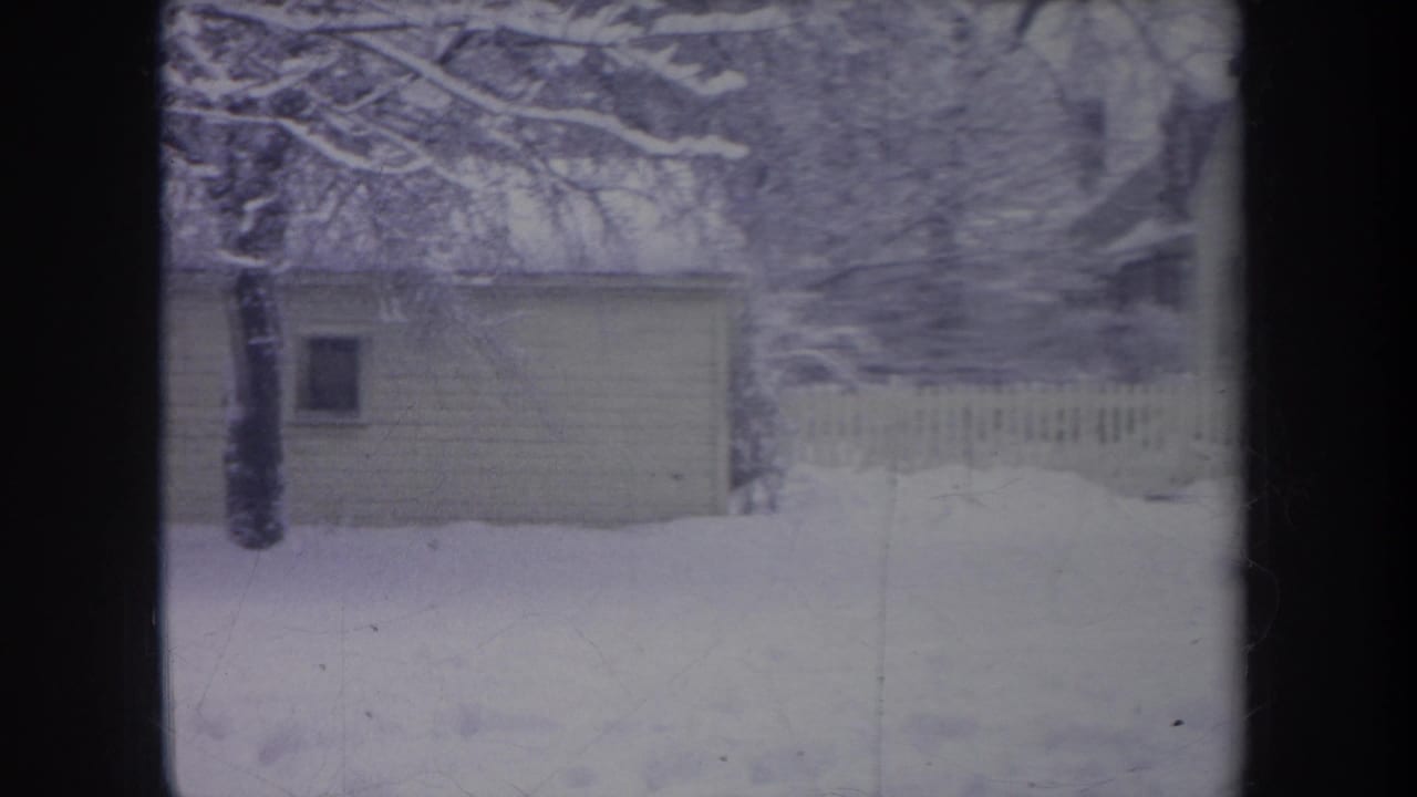 1968 Michigan home-movie view of a snowy yard with a small shed, tree, and pale picket fence in the background.