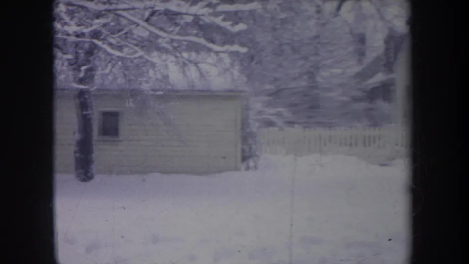 1968 Michigan home-movie view of a snowy yard with a small shed, tree, and pale picket fence in the background.