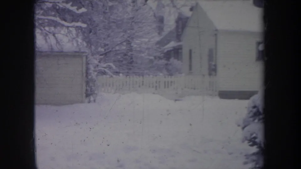 1968 Michigan 8mm frame showing a snowy residential yard, picket fence, shed edge, trees, and part of a modest house.