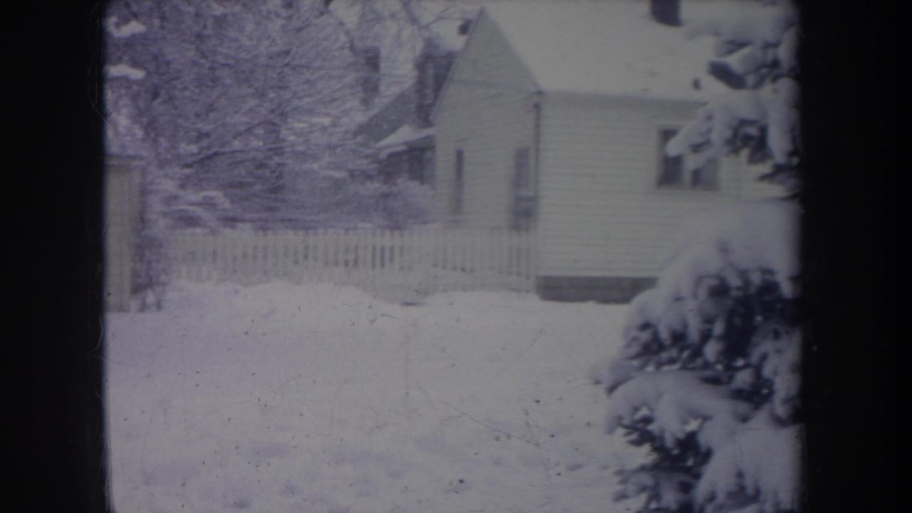 1968 Michigan 8mm frame showing a snowy suburban yard, white picket fence, house, bare trees, and heavy snow on evergreen branches.