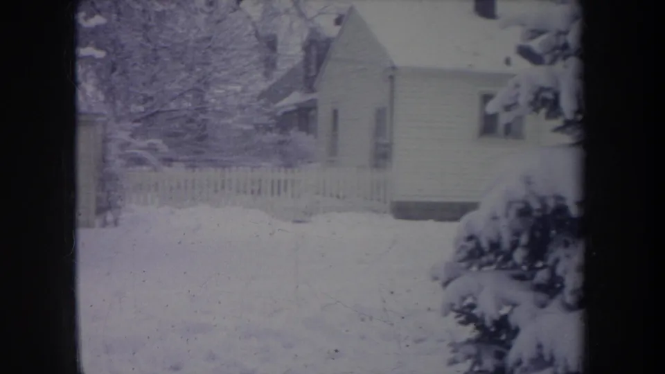 1968 Michigan 8mm frame showing a snowy suburban yard, white picket fence, house, bare trees, and heavy snow on evergreen branches.
