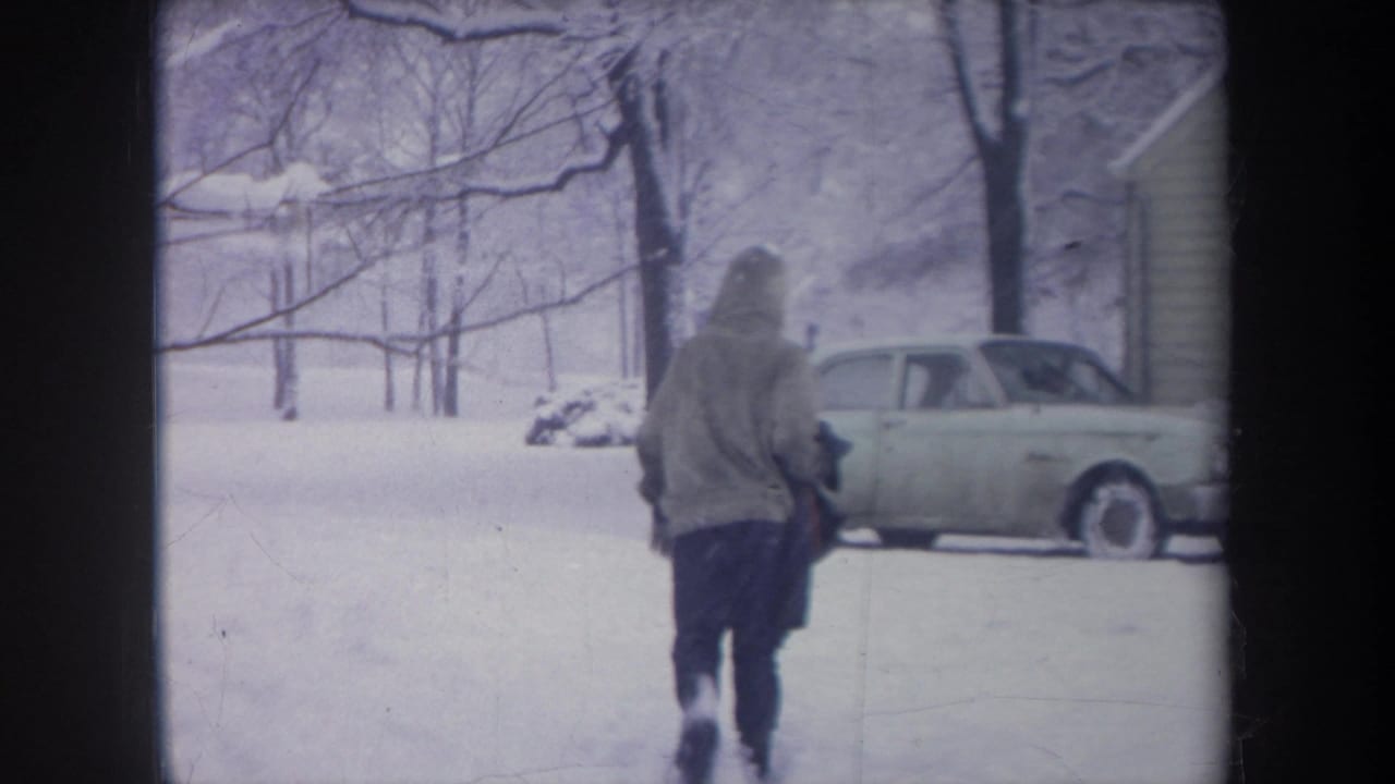 1968 Michigan 8mm frame showing a bundled person crossing deep snow toward a pale 1960s sedan parked near a house.