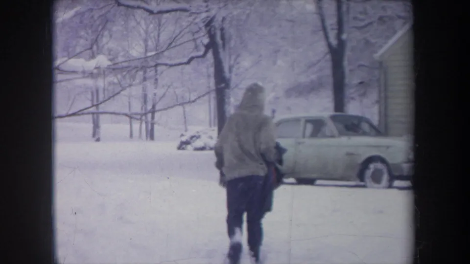 1968 Michigan 8mm frame showing a bundled person crossing deep snow toward a pale 1960s sedan parked near a house.