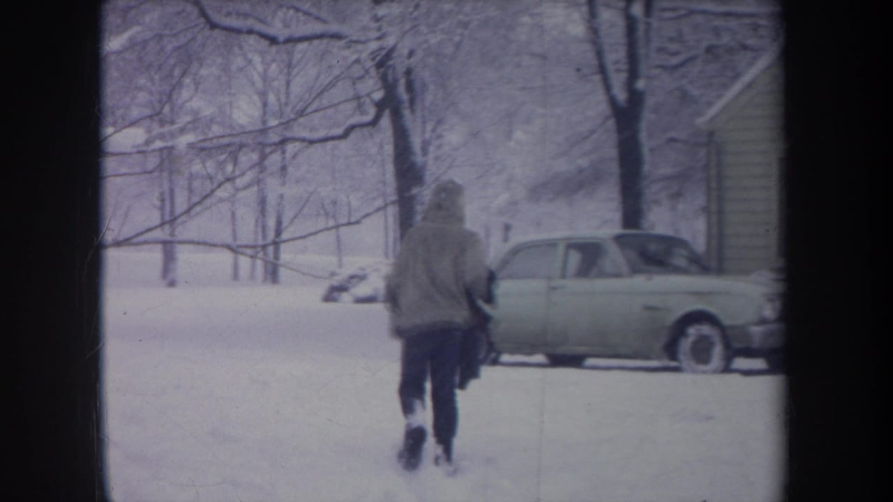 1968 Michigan 8mm frame of a hooded figure crossing a snowy residential yard toward a light-colored parked car.