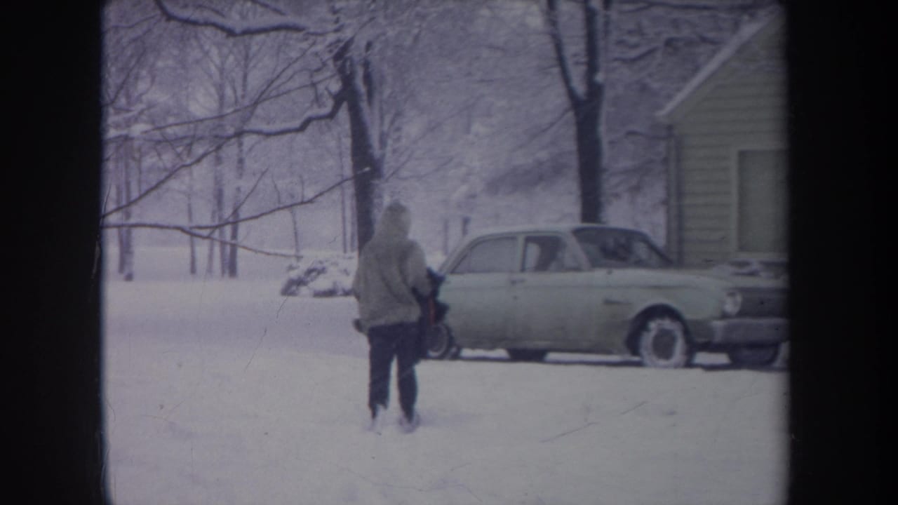 Hooded figure farther across the snowy yard beside a parked car.