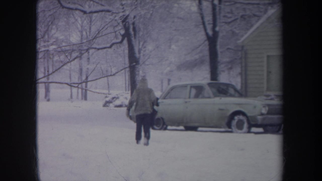 Rear view of a hooded figure in boots walking through deep snow toward a light-colored car in a Michigan residential scene.