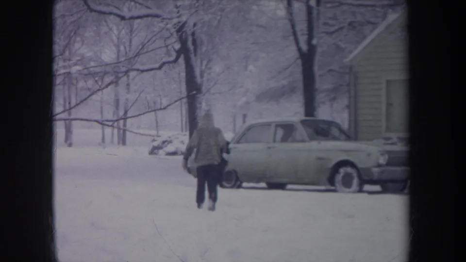 Rear view of a hooded figure in boots walking through deep snow toward a light-colored car in a Michigan residential scene.