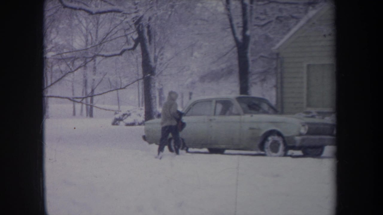 In 1968 Michigan home-movie footage, a hooded person approaches a light-colored sedan parked in deep snow near a house.