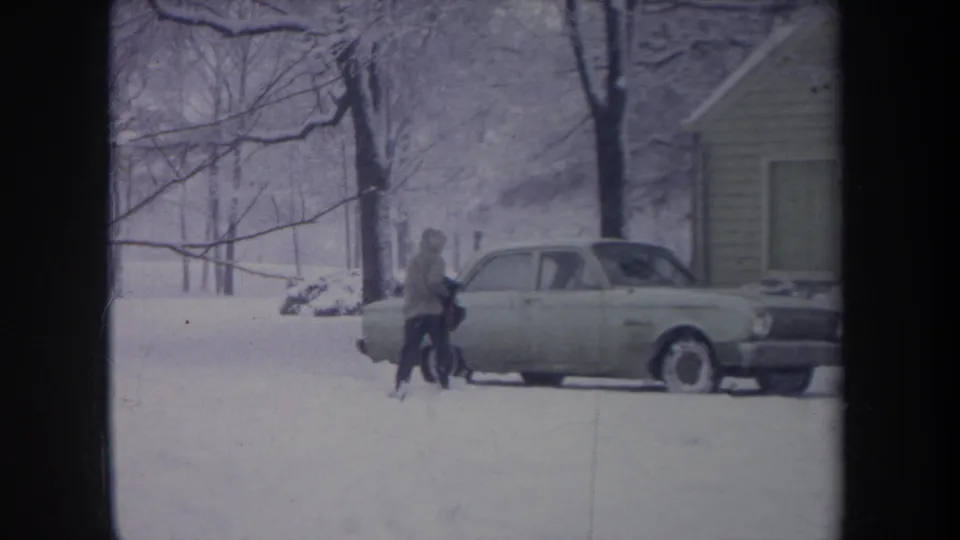 In 1968 Michigan home-movie footage, a hooded person approaches a light-colored sedan parked in deep snow near a house.