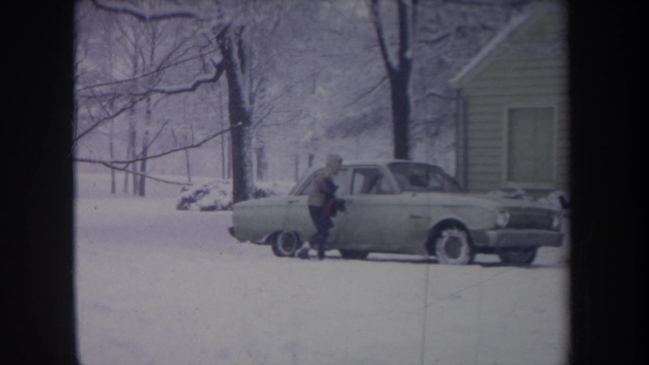 Hooded person stands near the rear passenger side of a snow-dusted sedan.