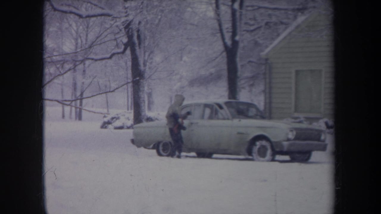 A hooded person pauses at the passenger side of the light-colored sedan during a snowy 1968 Michigan home-movie scene.