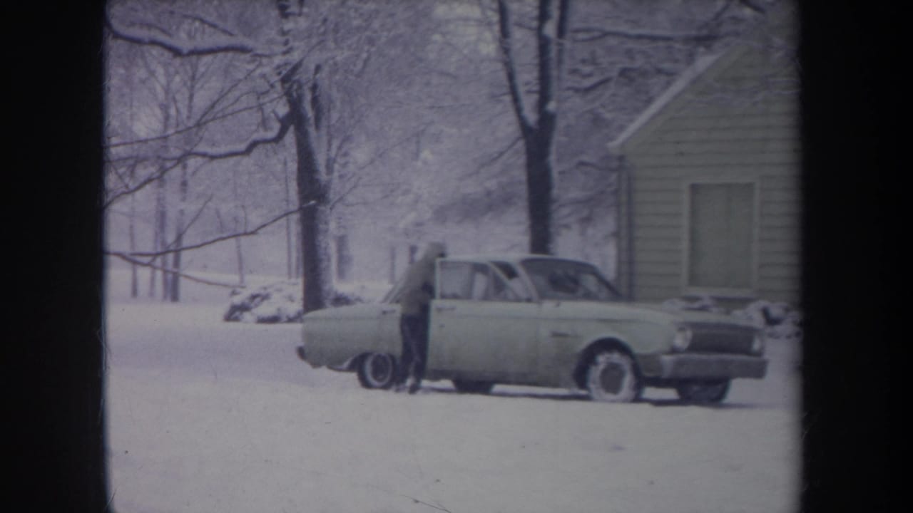 The passenger door of the light-colored sedan is open as the hooded person enters during the snowy Michigan winter scene.