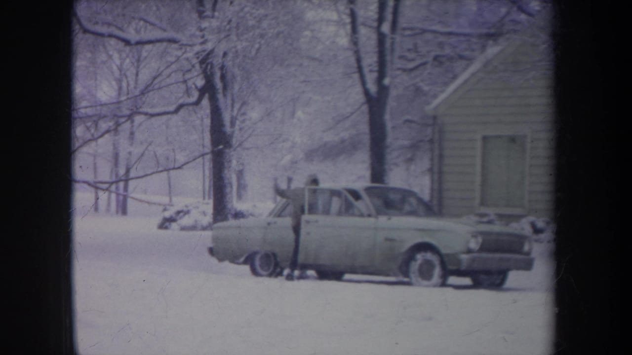 1968 Michigan 8mm frame showing a bundled person at the passenger side of a pale sedan beside a snow-covered house exterior.