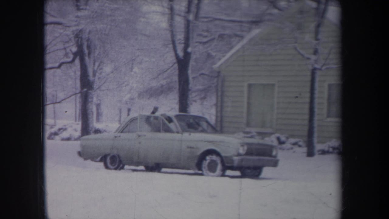 1968 Michigan home-movie frame of a pale sedan parked in a snowy yard near a modest house and bare winter trees.