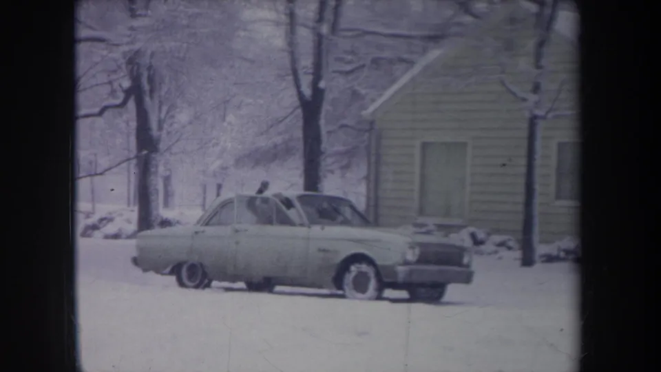 1968 Michigan home-movie frame of a pale sedan parked in a snowy yard near a modest house and bare winter trees.