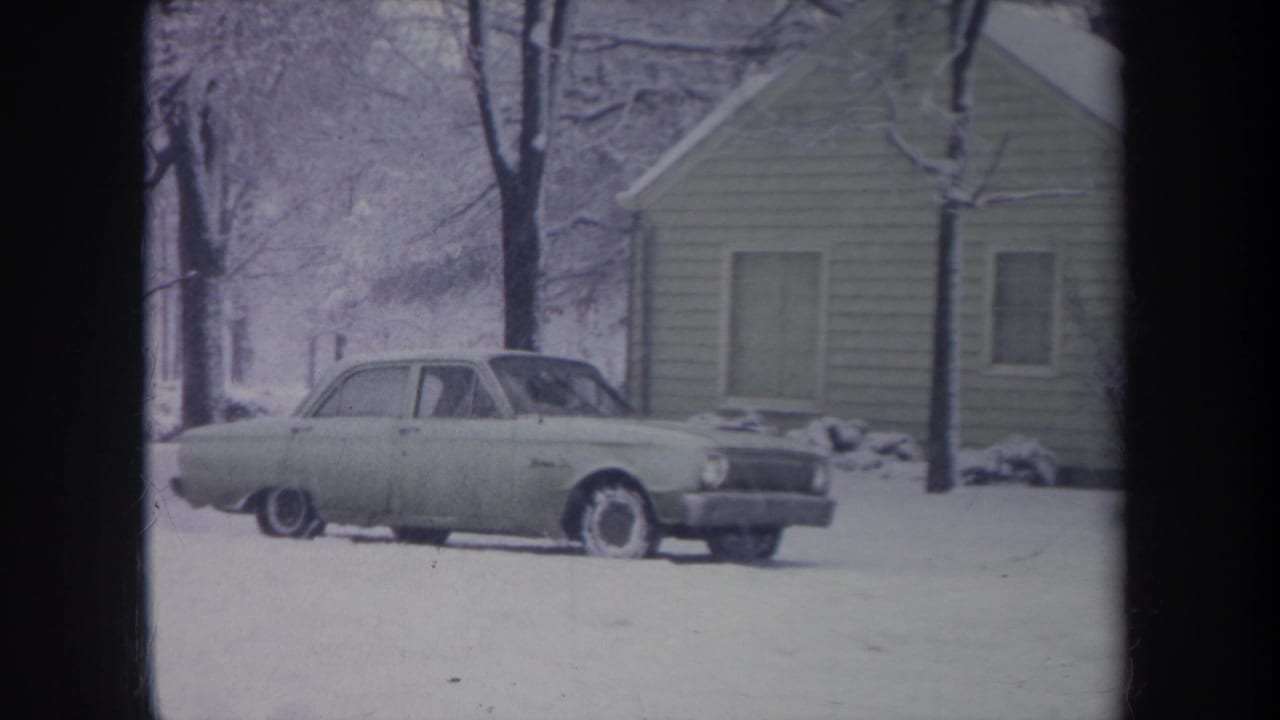 Final 1968 Michigan frame of the pale sedan parked in a snowy yard beside a modest house, with no clear person visible outside the car.