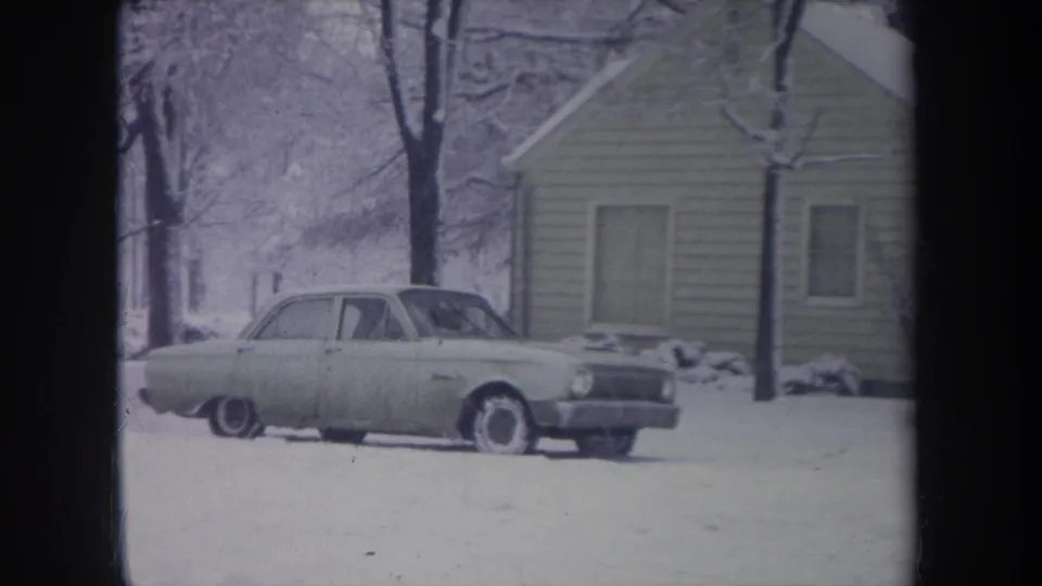 Final 1968 Michigan frame of the pale sedan parked in a snowy yard beside a modest house, with no clear person visible outside the car.