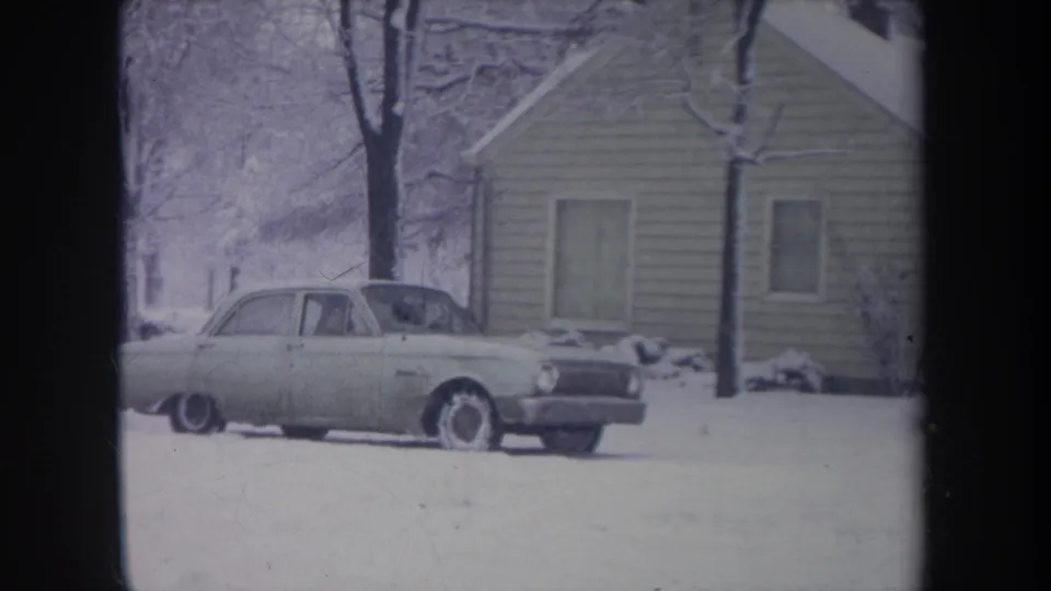 1968 Michigan home-movie frame of a light-colored sedan in a snowy residential yard beside a modest house.