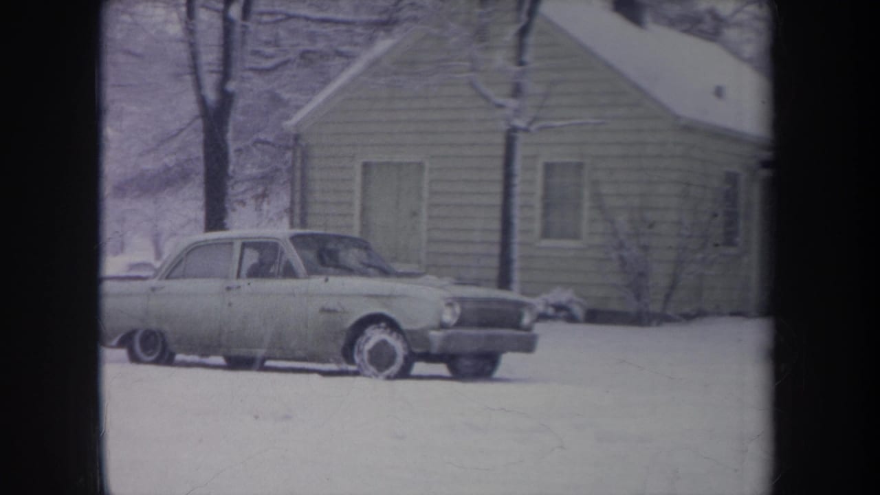 Light sedan in snow beside a small house, with a faint occupant visible through the window.