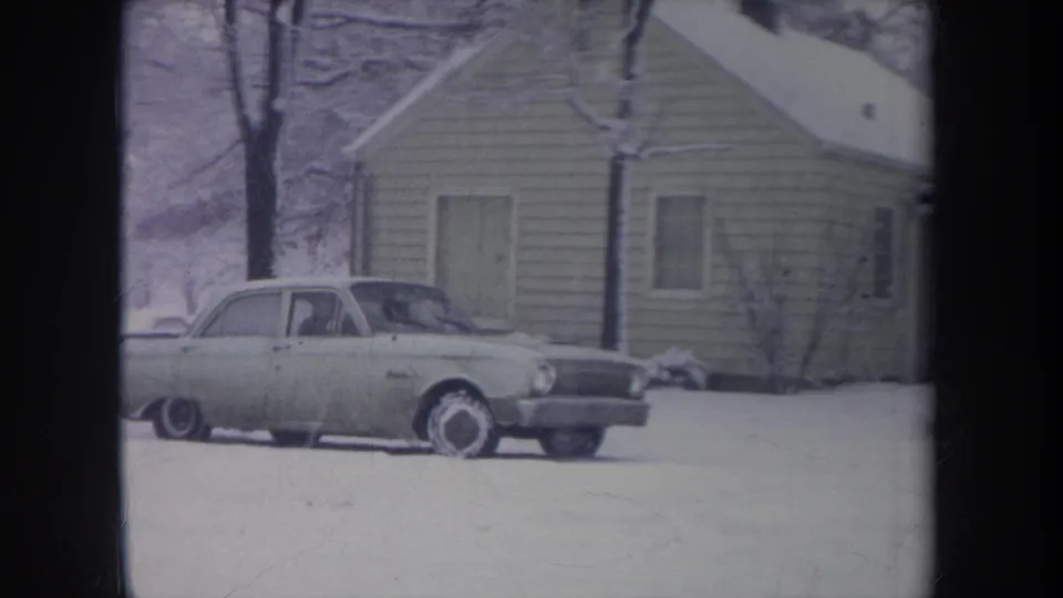 A light-colored older sedan passes slowly across a snowy Michigan yard, with a faint driver or occupant visible inside.