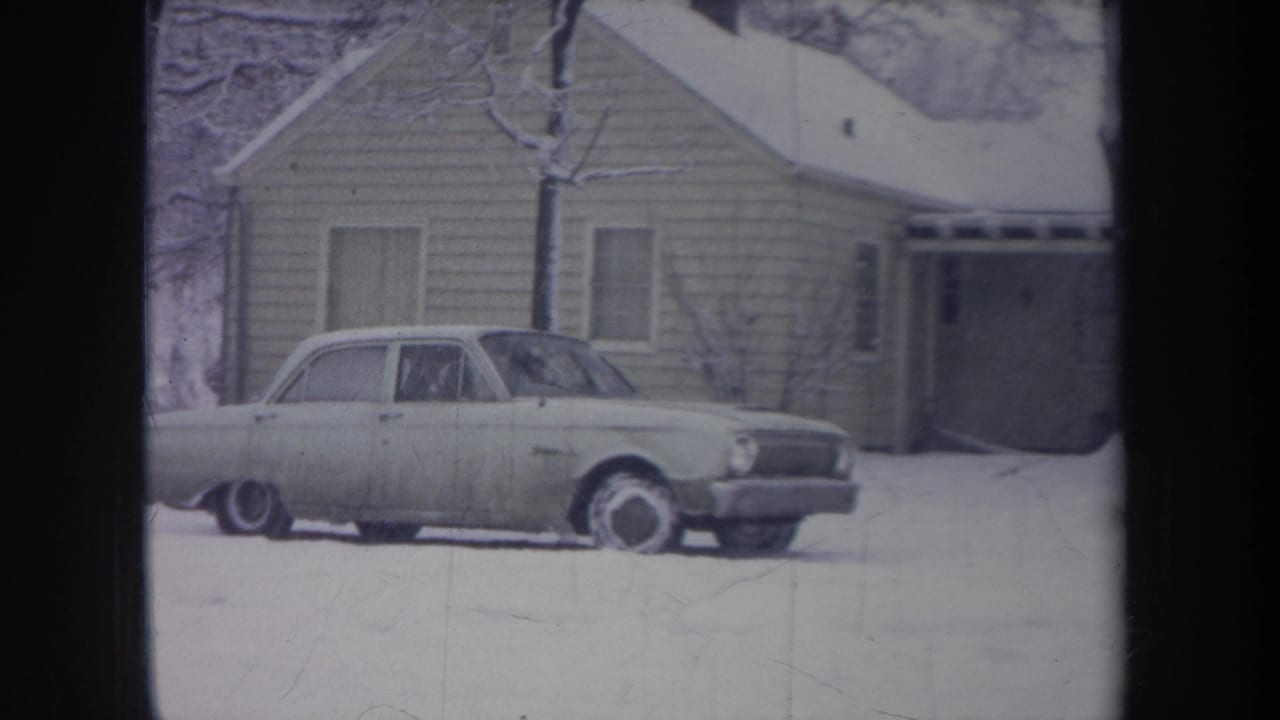 1968 Michigan frame showing an older light sedan progressing across a snow-covered yard near a porch-front house.