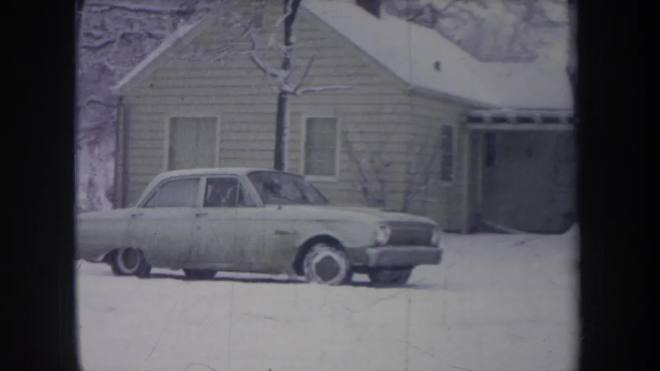 1968 Michigan frame showing an older light sedan progressing across a snow-covered yard near a porch-front house.