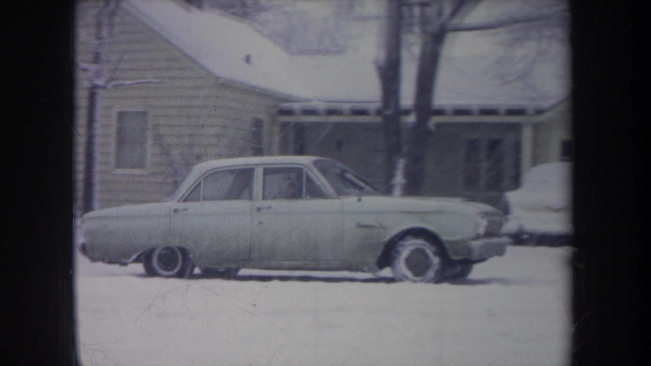 The light-colored sedan nears the right side of the snowy Michigan residential scene, with a porch and another vehicle partly visible.