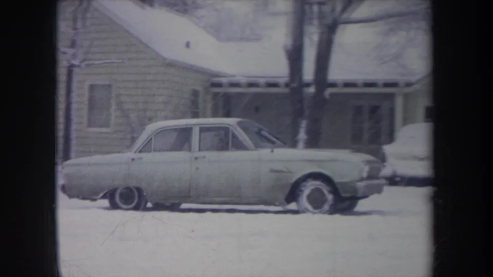 The light-colored sedan nears the right side of the snowy Michigan residential scene, with a porch and another vehicle partly visible.