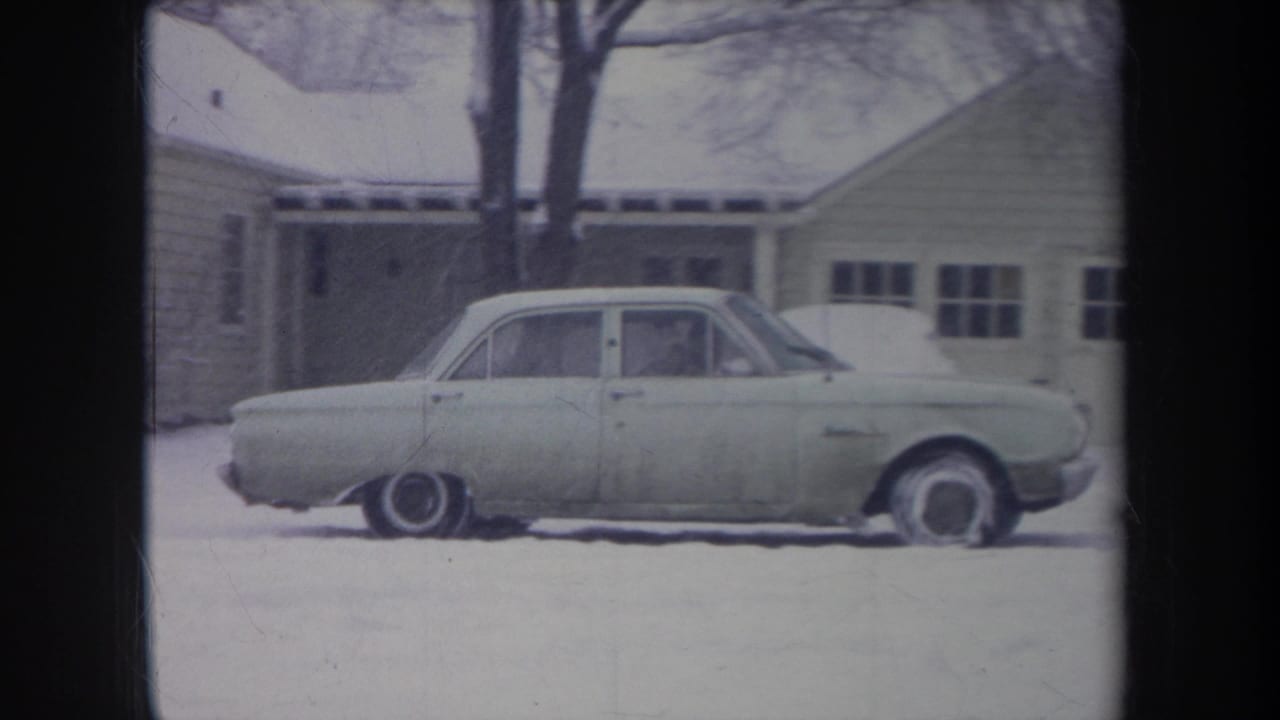 1968 Michigan home-movie frame showing a pale sedan in a snowy driveway or yard beside a modest house.