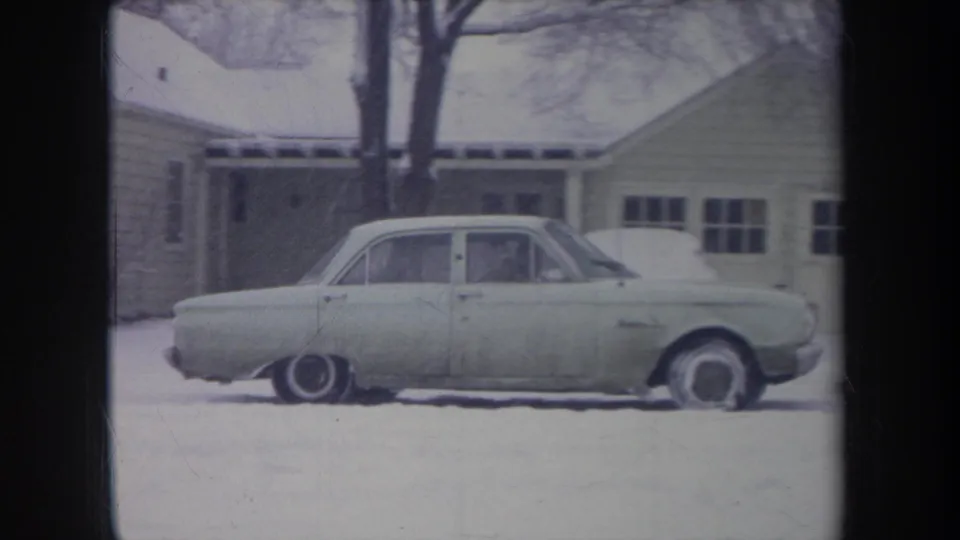 1968 Michigan home-movie frame showing a pale sedan in a snowy driveway or yard beside a modest house.