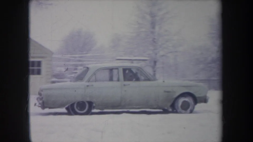 A pale 1960s sedan is shown side-on in snowy Michigan suburbia, with an indistinct occupant visible through the window.