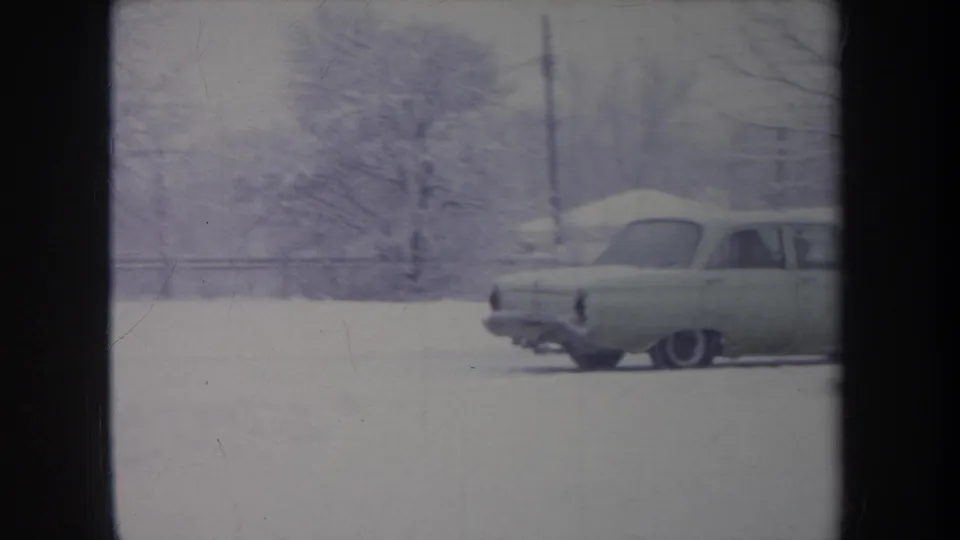 The light-colored sedan moves through snow in a residential Michigan setting, partly framed by winter trees.