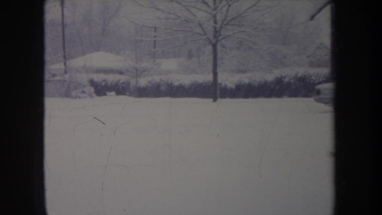 1968 metadata-supported 8mm view of a snow-covered Michigan residential street, framed by a dark window or door edge.