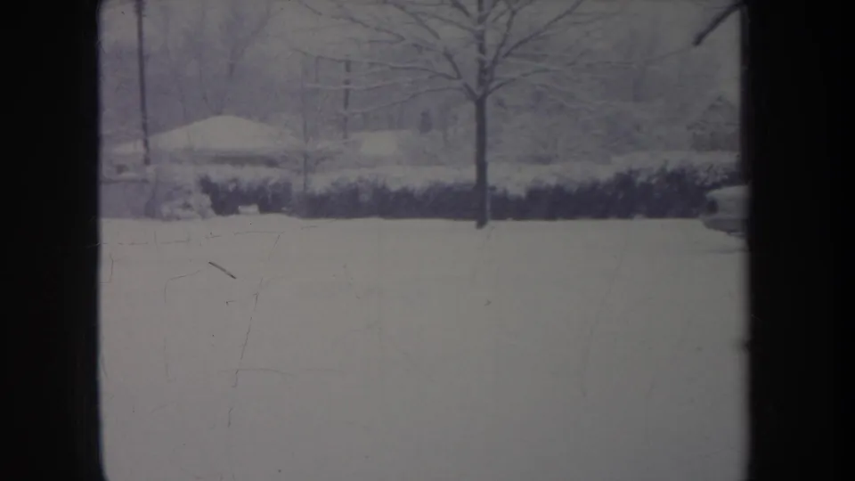 1968 metadata-supported 8mm view of a snow-covered Michigan residential street, framed by a dark window or door edge.