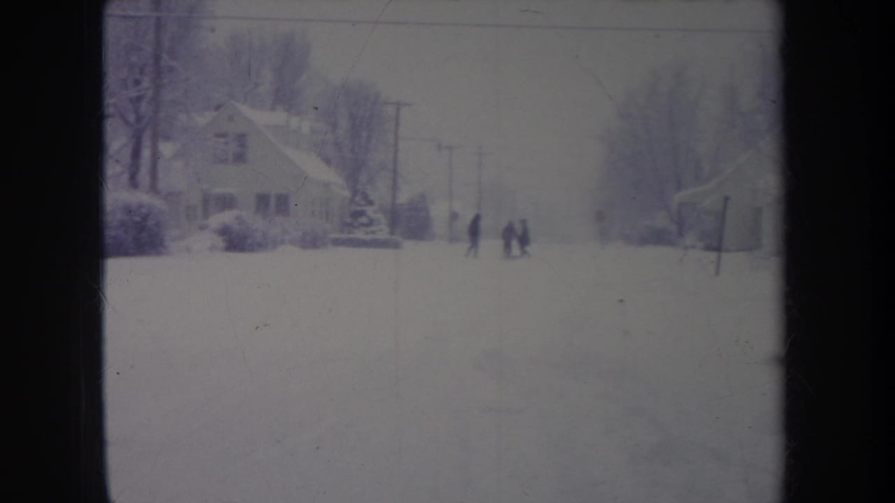 Snowy residential road with houses, utility poles, bare trees, and several distant figures in dark coats.