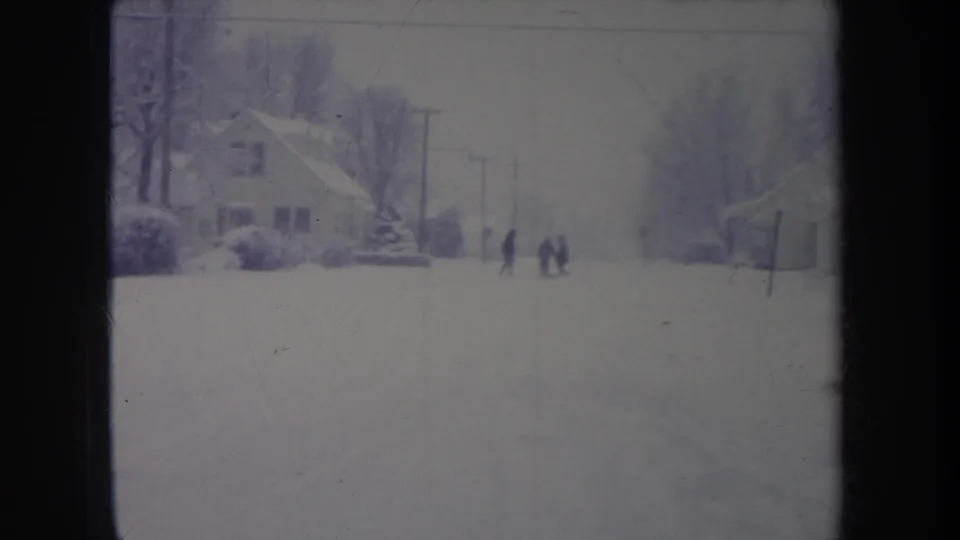 1968 metadata-supported 8mm frame of a snowy Michigan neighborhood with distant people walking or playing in the street.
