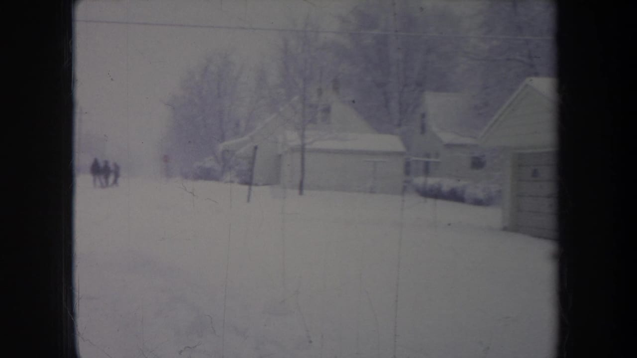 1968 Michigan home-movie frame showing a snow-covered residential area with garages, houses, bare trees, and distant figures.