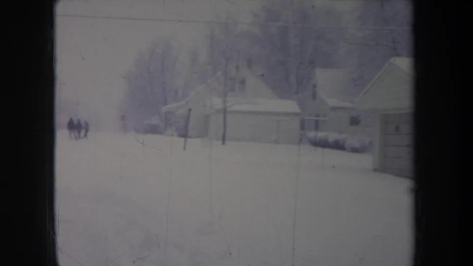 1968 Michigan home-movie frame showing a snow-covered residential area with garages, houses, bare trees, and distant figures.